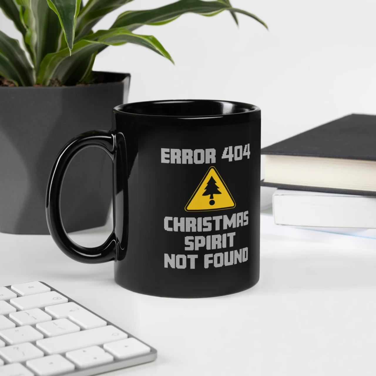 Black ceramic ERROR 404 Christmas Spirit Not Found Mug sitting on a desk next to a plant and books. The mug features a yellow warning triangle with a Christmas tree graphic and the text "ERROR 404 CHRISTMAS SPIRIT NOT FOUND." This is a funny Black Programmer Mug.