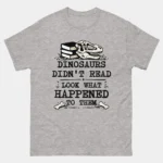 Sport grey Dinosaurs Didn't Read Funny Bookworm Tee featuring a dinosaur skull resting on stacked books with crossed bones and bold text reading Dinosaurs Didn't Read Look What Happened To Them, funny teacher and reading lover graphic tee